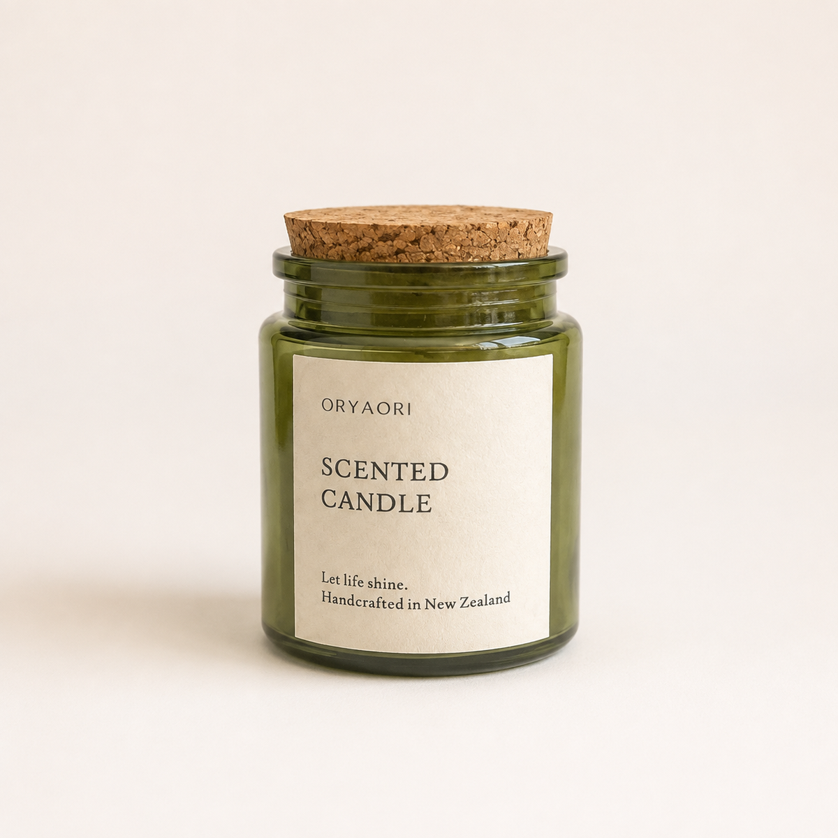 An Oryaori scented candle in a dark green glass jar with a natural cork lid and a cream-colored paper label, set against a plain off-white background.