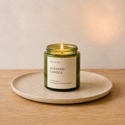 A green glass jar candle with a white label reading 'ORYAORI SCENTED CANDLE' sits lit on a round beige ceramic tray over a light wood surface against a warm beige wall.