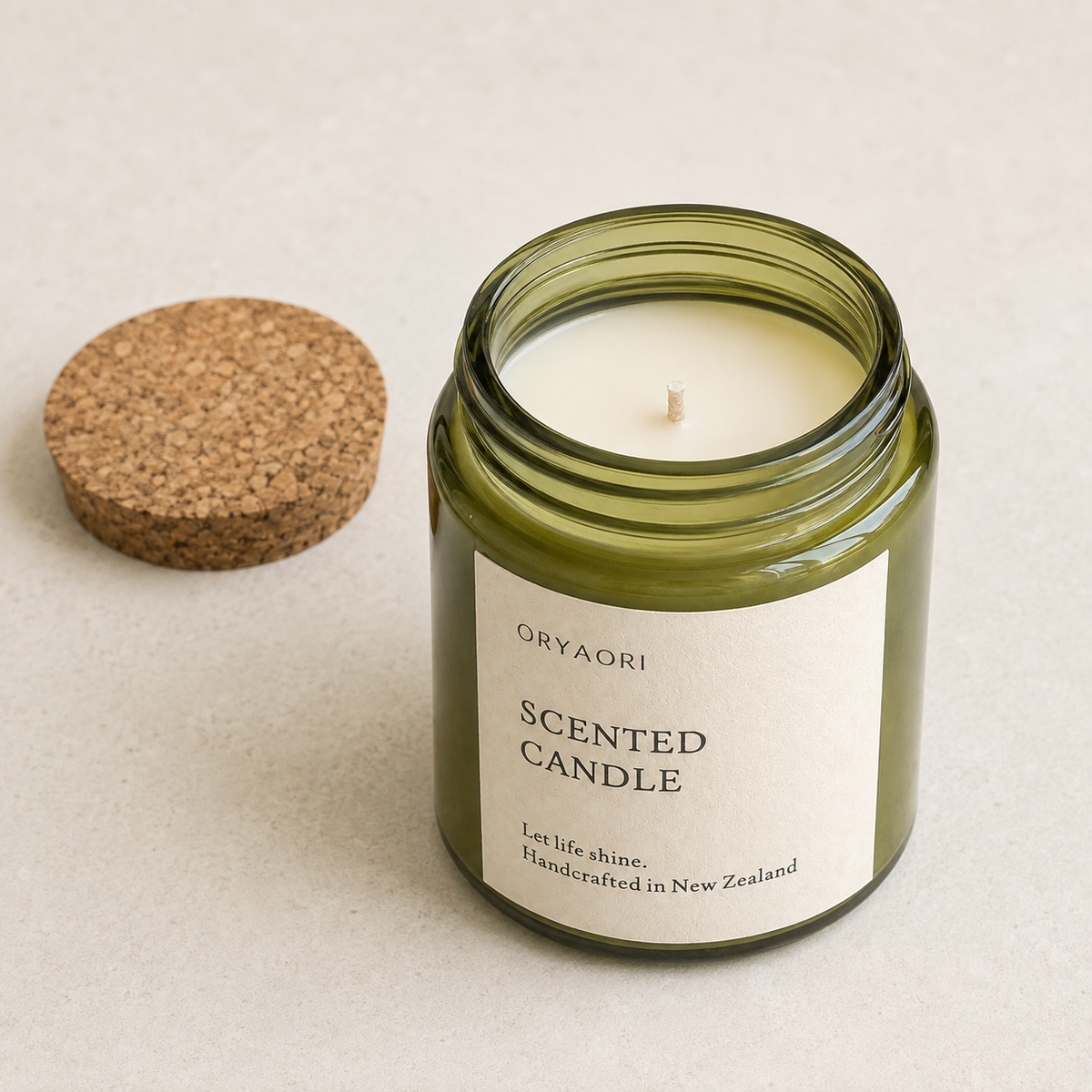An open scented candle in a translucent olive green glass jar with a cork lid resting beside it on a light beige textured surface. The label reads 'ORYAORI SCENTED CANDLE'.