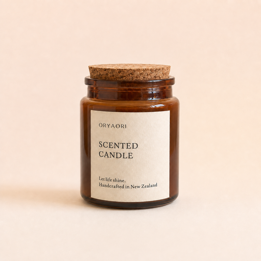 An amber glass jar candle with a cork lid and a cream paper label reading 'ORYAORI SCENTED CANDLE' on a warm beige background.