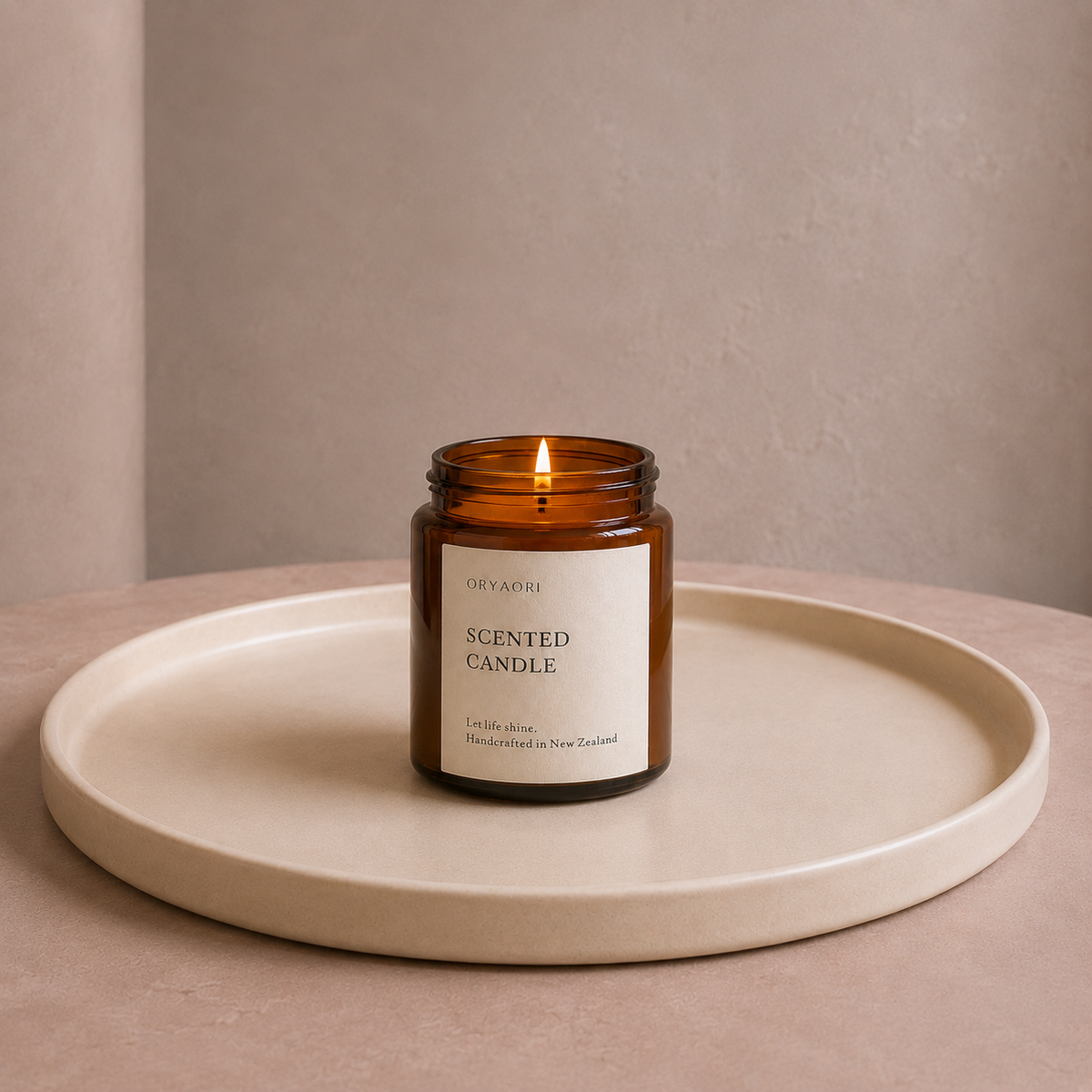 A lit amber glass scented candle with a cream label sits centered on a round beige tray against a soft neutral wall.