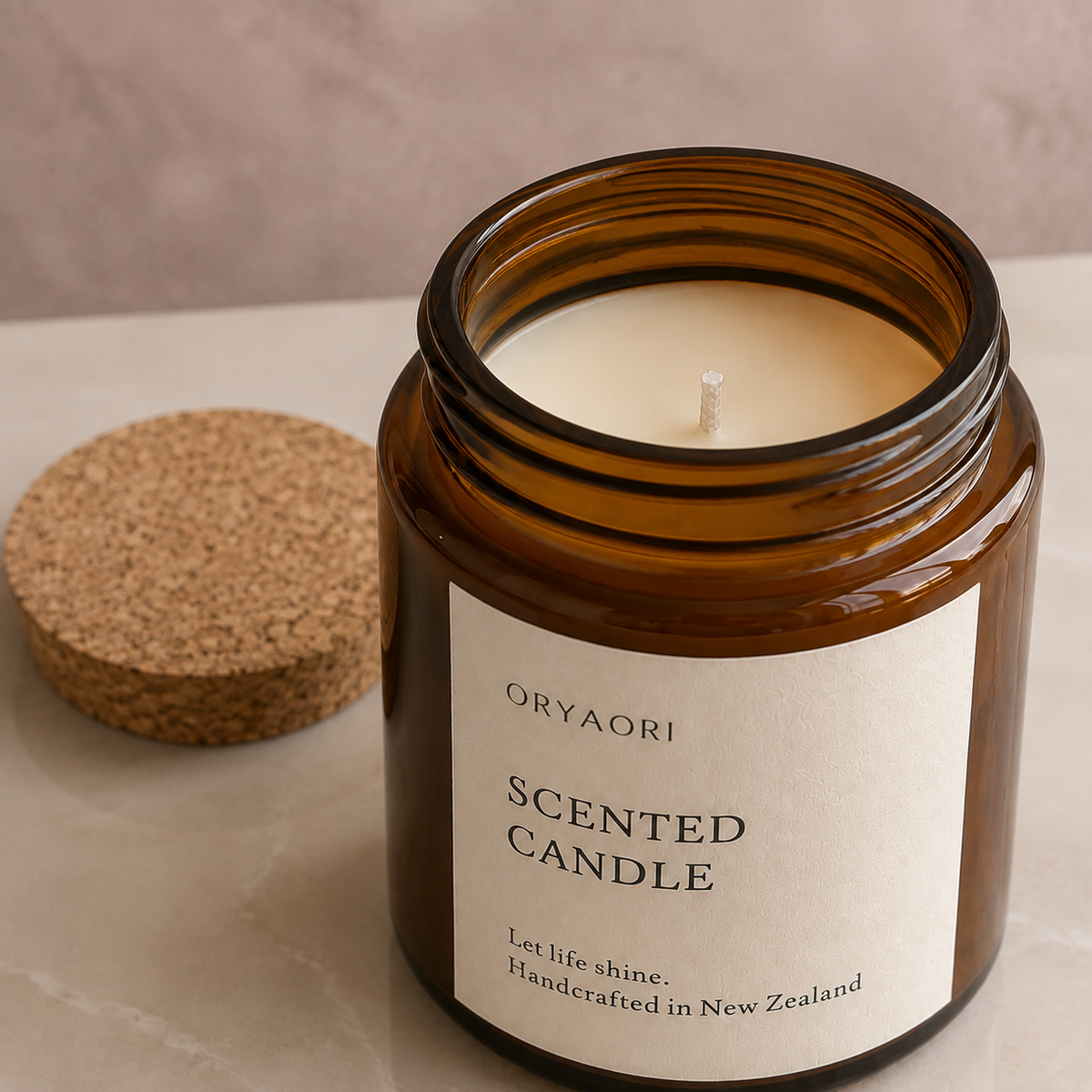 An amber glass jar candle with a cream-colored wax and a white label reading 'ORYAORI SCENTED CANDLE' sits on a light stone surface next to its cork lid.