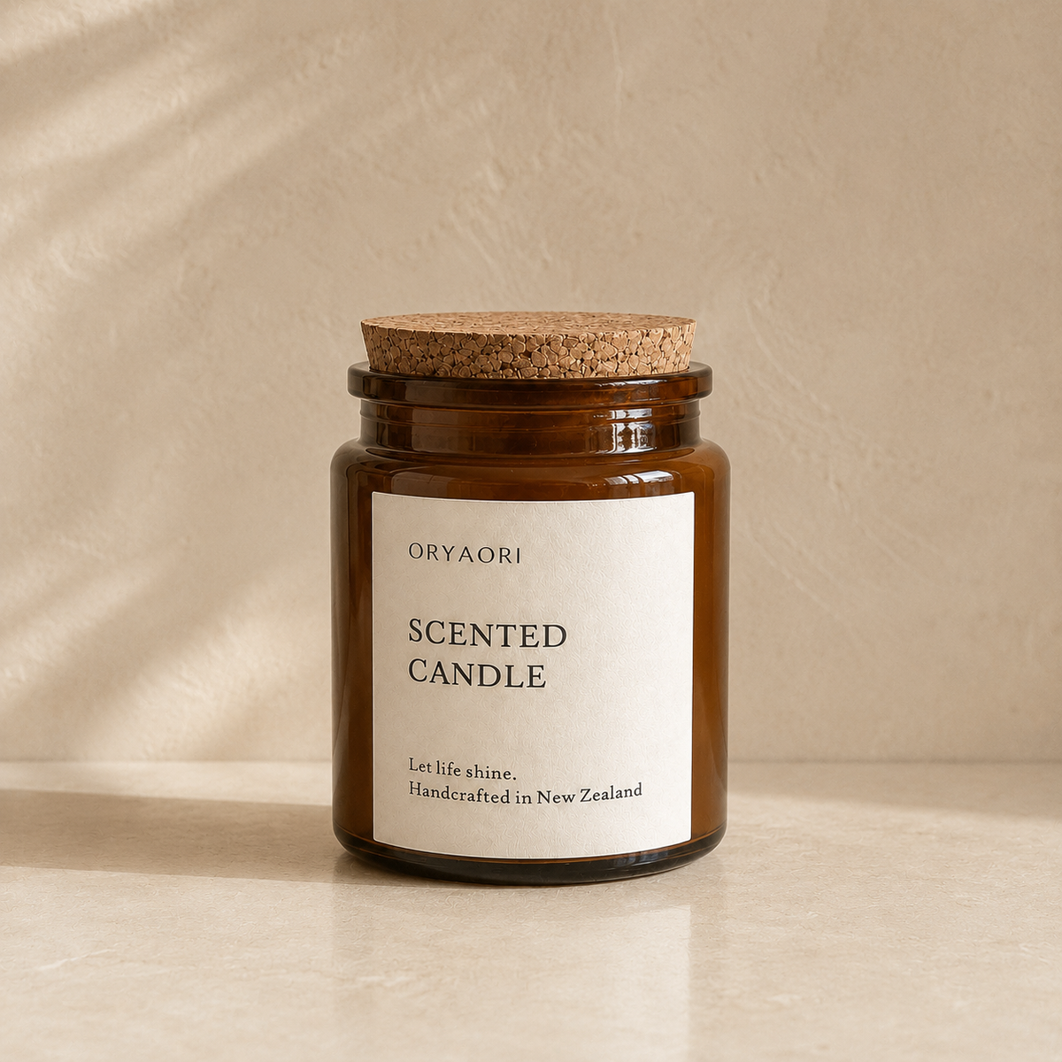 An amber glass jar candle with a cork lid and a cream paper label reading 'ORYAORI SCENTED CANDLE' on a warm beige surface and backdrop.
