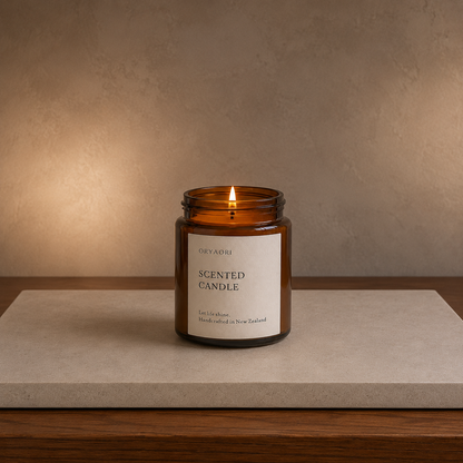 An amber glass jar candle with a white label reading 'Oryaori Scented Candle' sits lit on a light stone slab over a wooden surface against a warm beige wall.