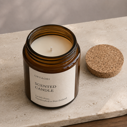 An amber glass scented candle jar with a cream label reading ORYAORI SCENTED CANDLE, shown open with the cork lid resting beside it on a light stone surface.