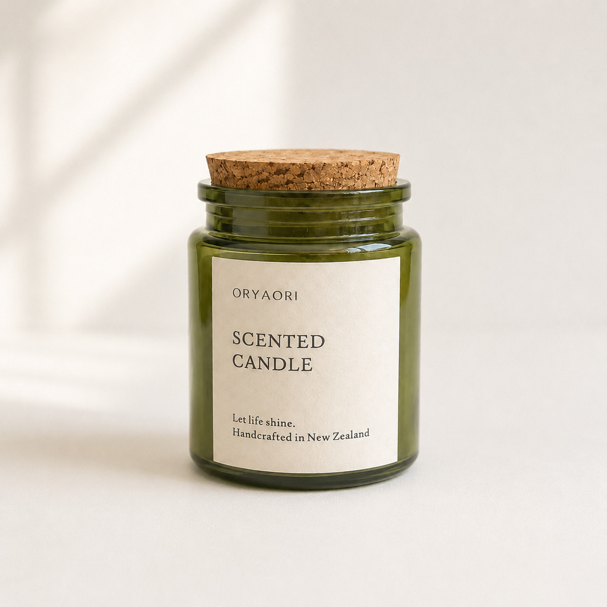 An Oryaori scented candle in a dark green glass jar with a natural cork lid and a minimalist white label, set against a clean white background with soft window shadow patterns.