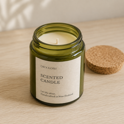 An open green glass scented candle jar with a cream label reading ORYAORI SCENTED CANDLE, set on a light wood surface with a cork lid beside it.