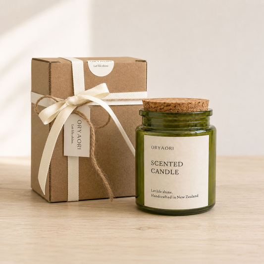 An Oryaori scented candle in an olive green glass jar with a cork stopper, shown next to its brown kraft paper gift box tied with a cream ribbon on a light wood surface.