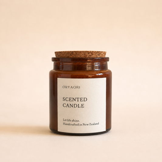 An amber glass scented candle jar with a cork lid and a minimalist white label reading 'ORYAORI SCENTED CANDLE' on a warm beige background.