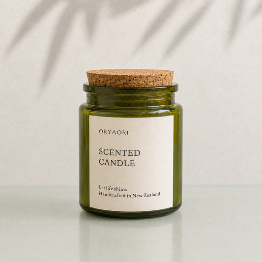 An Oryaori scented candle in an olive green glass jar with a natural cork lid and a cream paper label, set on a light gray surface with soft leaf shadows on a textured light gray wall.