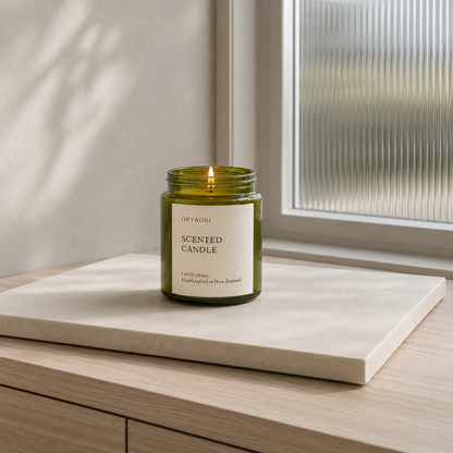 A green glass jar candle with a white label reading 'ORYAORI SCENTED CANDLE' sits lit on a light stone tray atop a wooden surface near a window.
