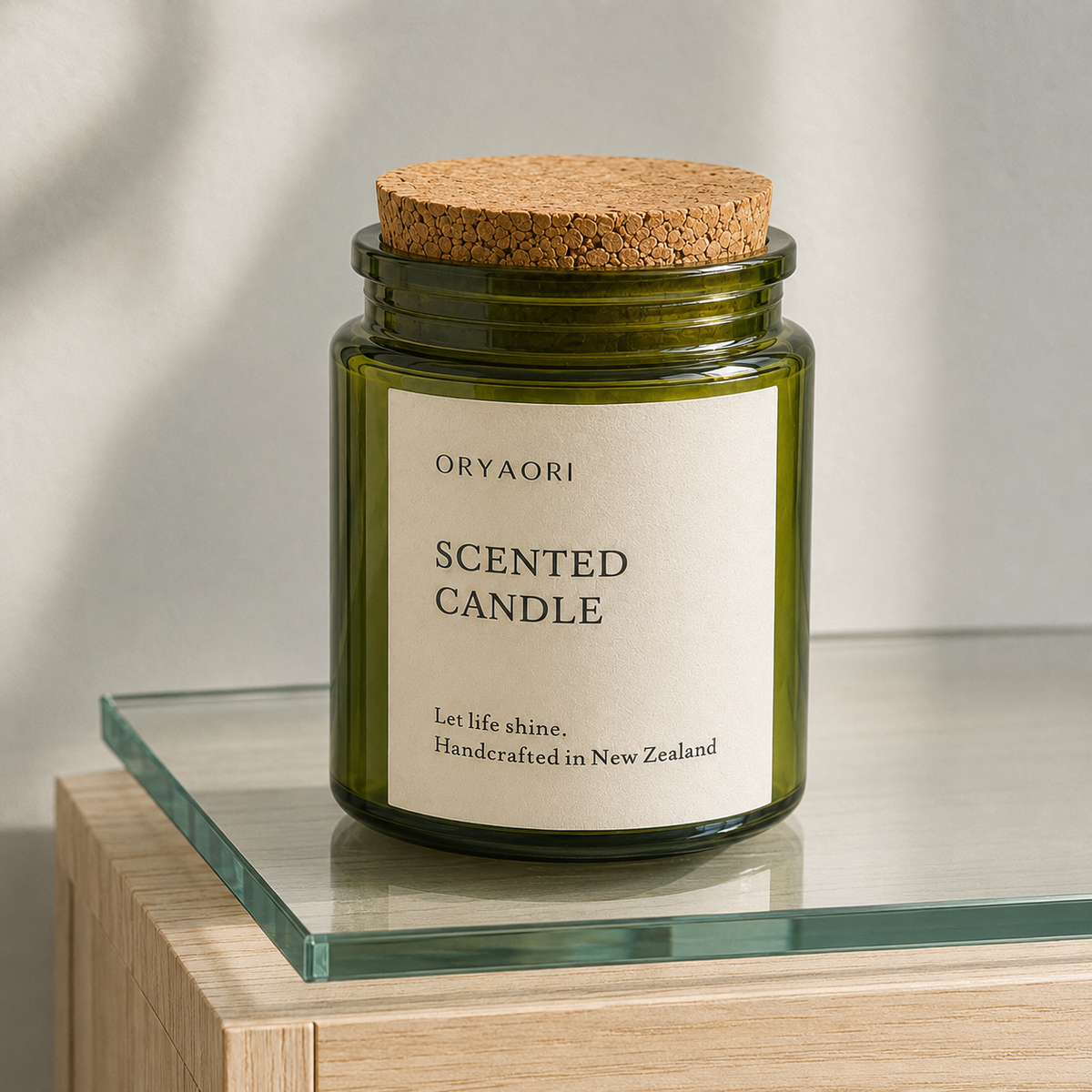 An Oryaori scented candle in an olive green glass jar with a cork lid, resting on a glass shelf of a light wood table against a neutral background.